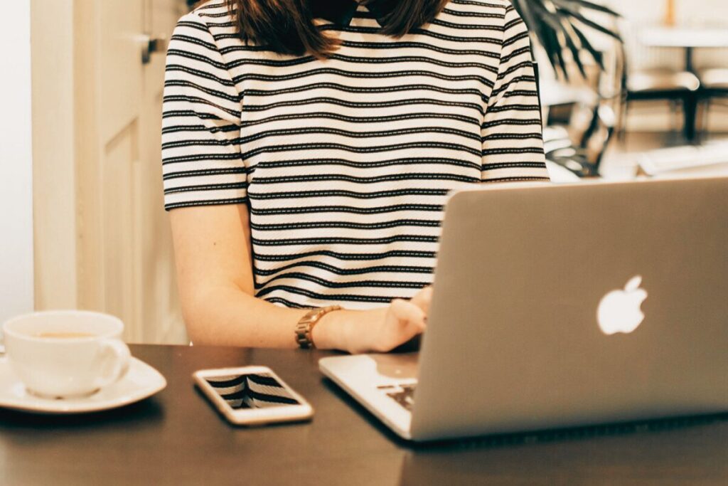 女性がMacBookのノートパソコンを操作しているのを肩から下の正面から写した画像。女性の右手にはスマホと白いコーヒーカップ&ソーサーが置いている