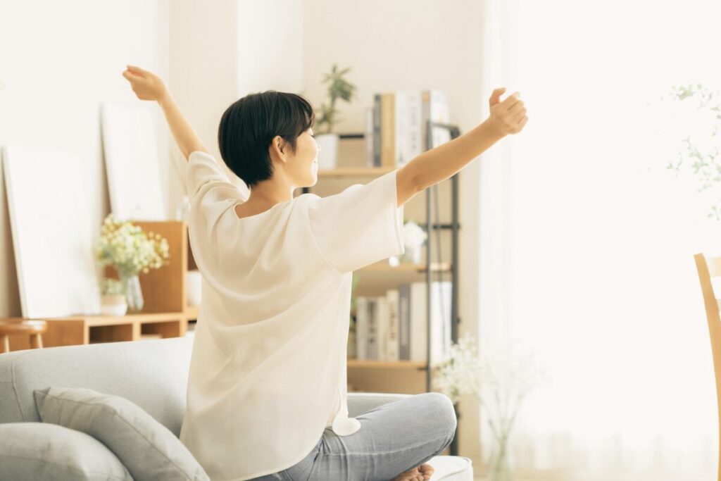 明るい部屋でショートカットの女性が窓の方を向いて両手を広げている様子を斜め後ろから写した写真。