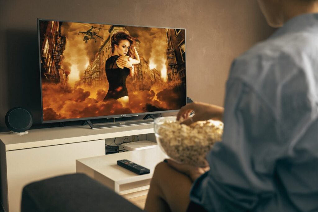 ポップコーンを食べながらテレビを見ている女性を左斜後ろから捉えている。海外の美しい女性が戦っているような映像を見ている。