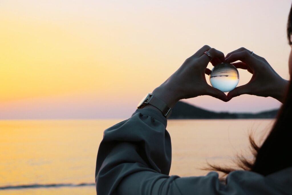 夕焼けの海に向かって女性が手をハート型にしながら水晶のようなガラスの玉を持って眺めている。